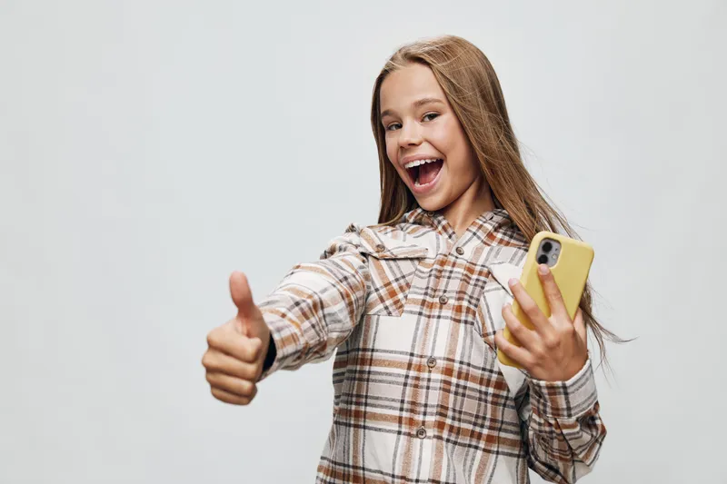 Girl smiling with thumb up and smartphone in hand wearing checked shirt standing on light background. Positive expression, technology and happiness, communication and youth lifestyle.