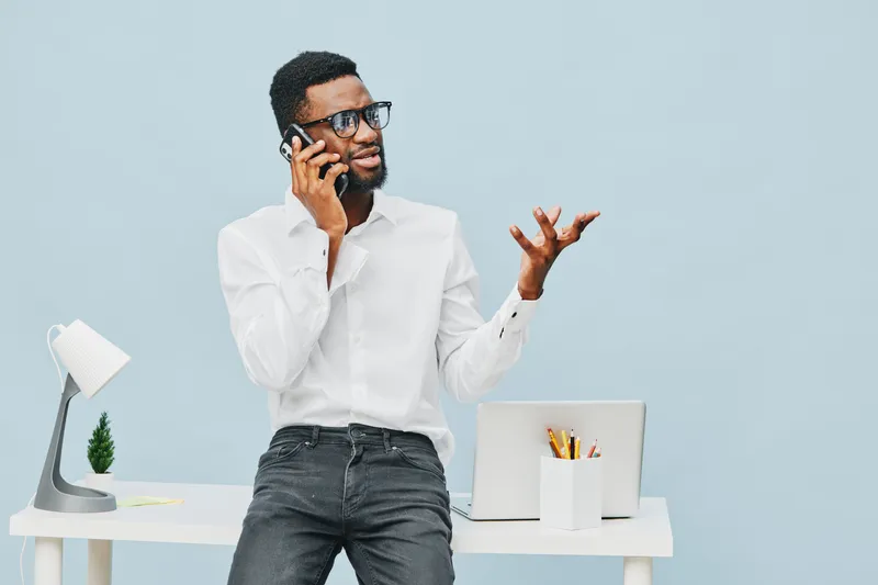 Modern young man with glasses, beard, casual white shirt, talking on smartphone while standing by white desk with laptop and stationery, light blue background, indoor workspace. People lifestyle conce