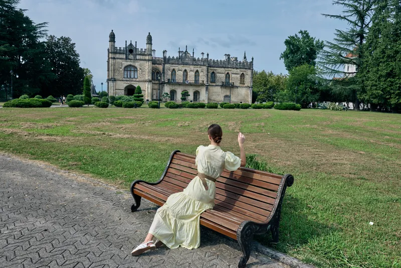 Woman in a vintage dress sits on a bench in a park with a historic castle and ornate architecture in the background, exuding leisure and timeless outdoor photography.