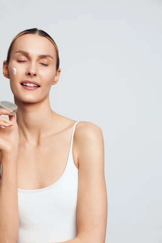Bright, minimalist portrait of a woman applying moisturizer to her face, eyes closed, serene smile, soft studio light, clean aesthetic, healthy glow, fresh skincare moment