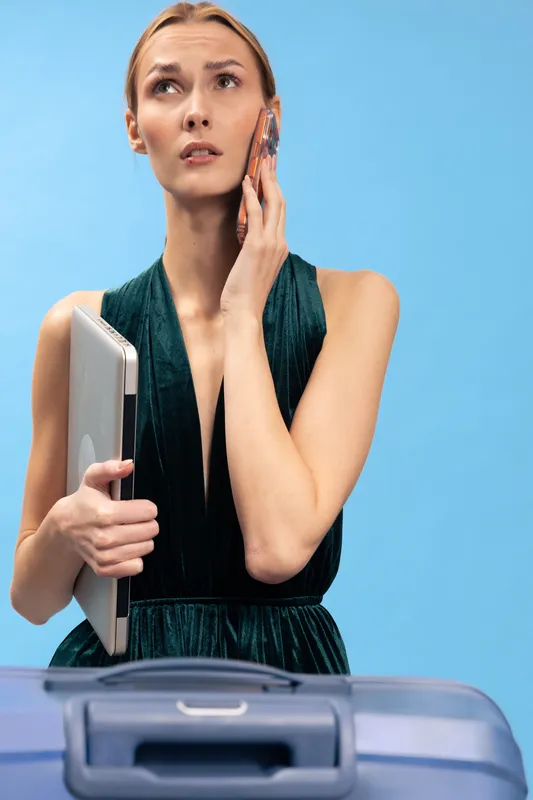 Concerned woman holds laptop and talks on phone near suitcase on blue background. Concept for travel planning, business trip, stress, communication, and technology usage.