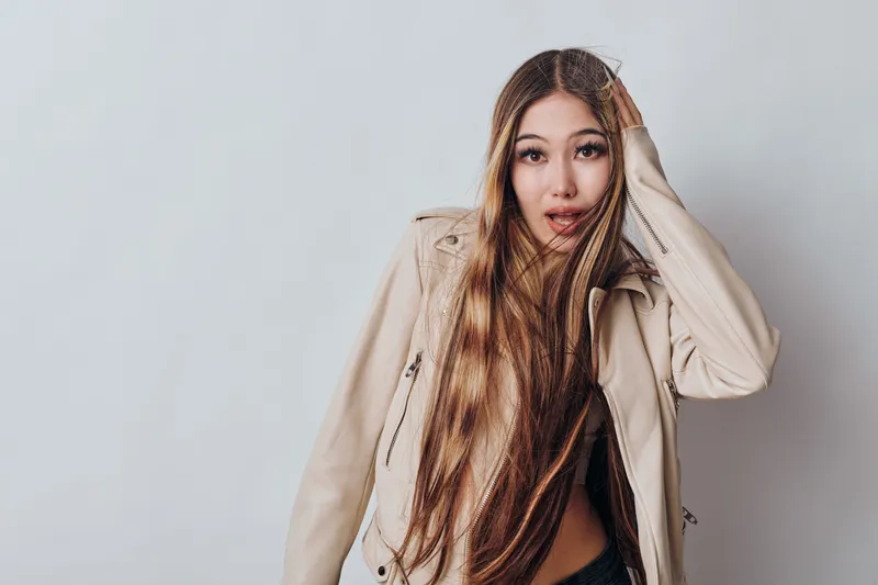 Young woman with long braided hair wearing beige jacket looking surprised or curious with hand on head against plain light background in studio portrait photography.