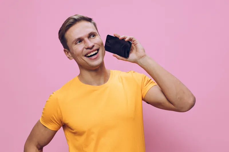 Smiling man talking smartphone vibrant orange t-shirt pink background casual communication digital nomad concept
