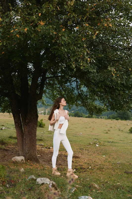 Young woman with curly hair stands thoughtfully under a large tree in a natural field wearing white pants and a white top, enjoying a peaceful moment outdoors.