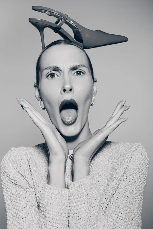Surprised Caucasian woman with pale skin wearing knitted sweater holds high heel shoe on head. Concept for fashion, emotion, and creativity in studio.