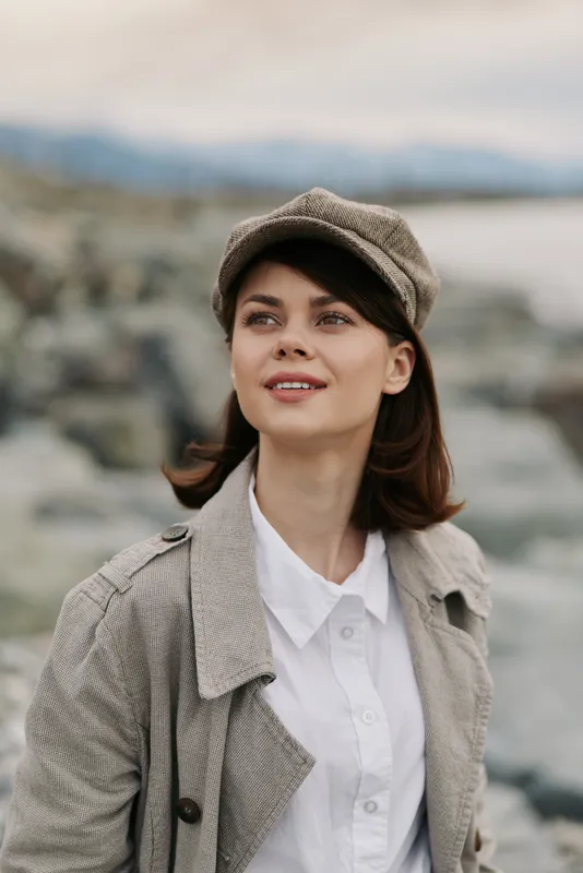Young woman wearing a patterned coat and cap smiles outdoors near a rocky shore. Soft natural light highlights her face, creating a calm and confident mood in a casual autumn setting.
