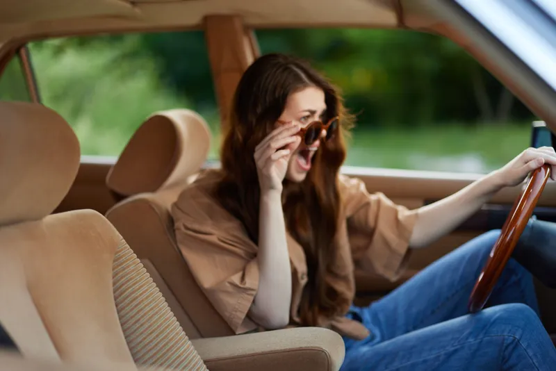 A young woman with long brown hair, wearing a casual brown oversized shirt, joyfully driving a vintage car on a sunny day in a green environment.