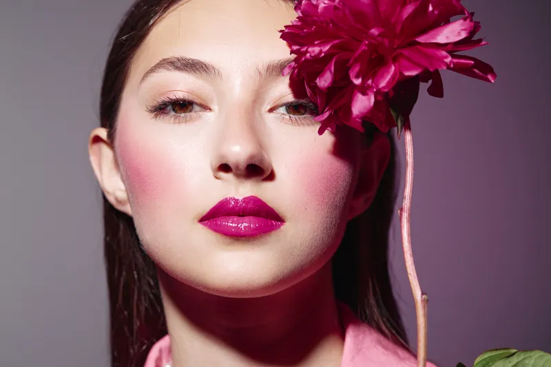 Young woman poses with bright pink makeup and lips holding dark pink flower in front of face on pink gradient background. Beauty creative concept.
