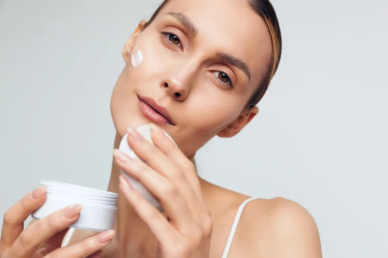 Young woman with clear skin applying white face cream on cheek. Close-up portrait with neutral background. Concept of skincare, beauty routine, hydration, and self-care.