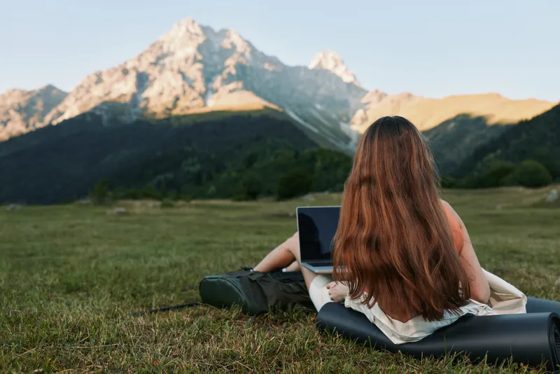 A cheerful professional sits outdoors with a laptop in a scenic mountain setting, smiling and focused, projecting productivity and creativity in nature.