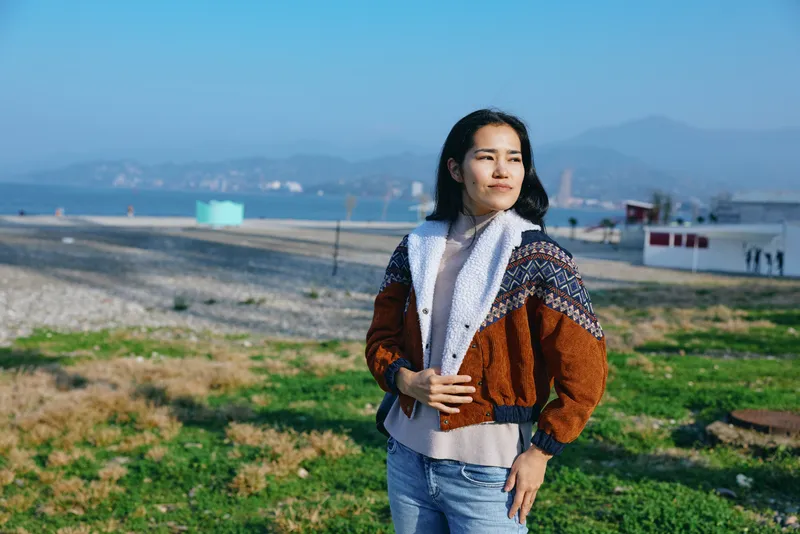 Woman standing outdoors in a knit sweater and jacket, overlooking a coastal field with distant hills, clear sky, and calm scenery for a relaxed lifestyle image.