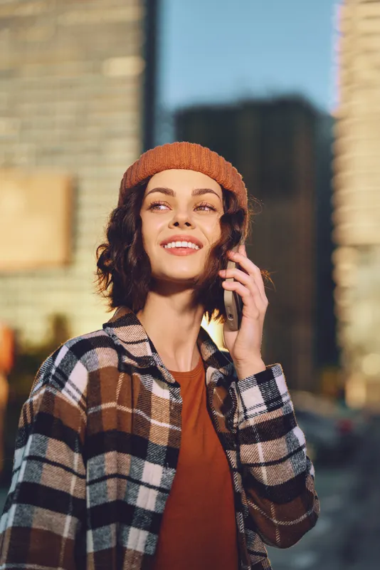 Woman phone smile urban beanie sunlight candid lifestyle portrait of a smiling woman on phone in city at golden hour, plaid jacket and warm glow emphasizing authenticity, mindful living, emotional