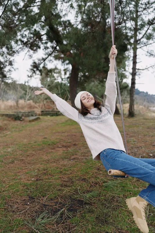 Woman on a rustic swing in an outdoor park, arms raised with a joyful smile, wearing a casual sweater and jeans, natural scenery with trees and grass, carefree moment in fresh air