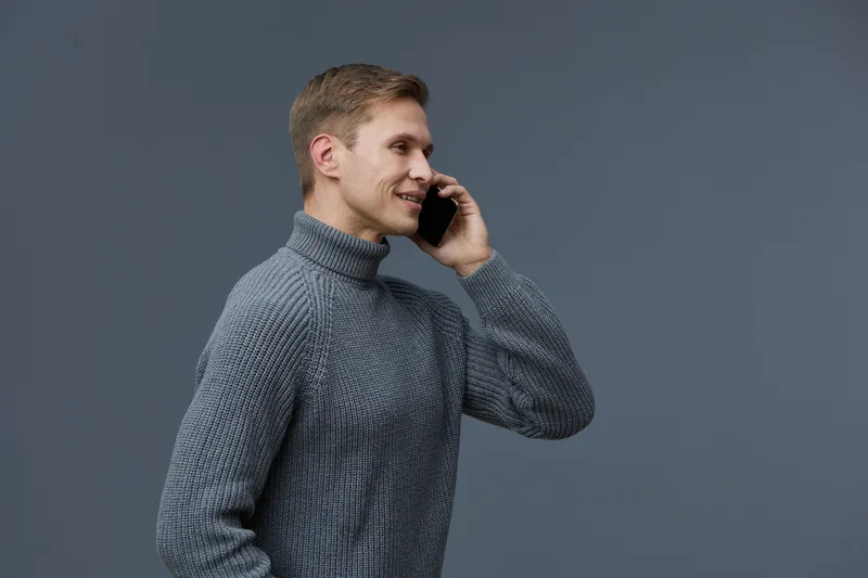 Professional young man with blonde hair wearing cozy gray turtleneck sweater smiling while talking on mobile phone, isolated on plain gray background, studio portrait, lifestyle concept, casual fashio
