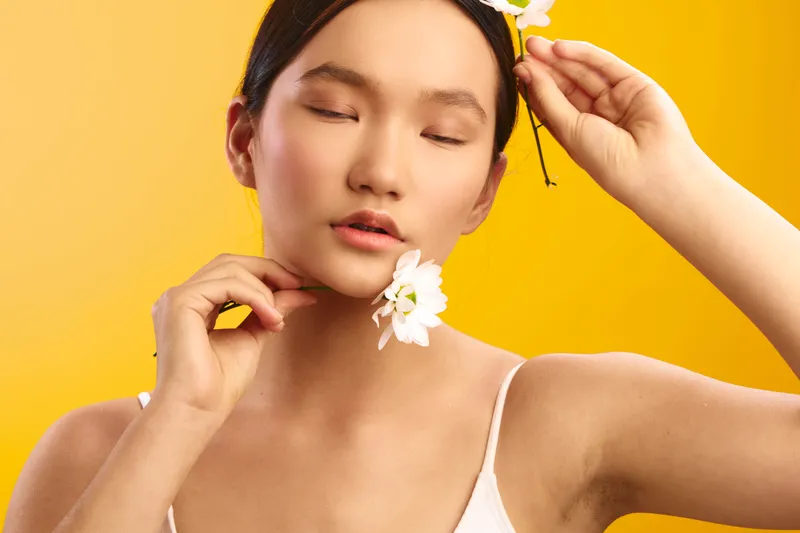 asian woman wearing a white top stands against a bright yellow backdrop, eyes closed as she adjusts a flower behind her ear, exuding calm and graceful beauty.