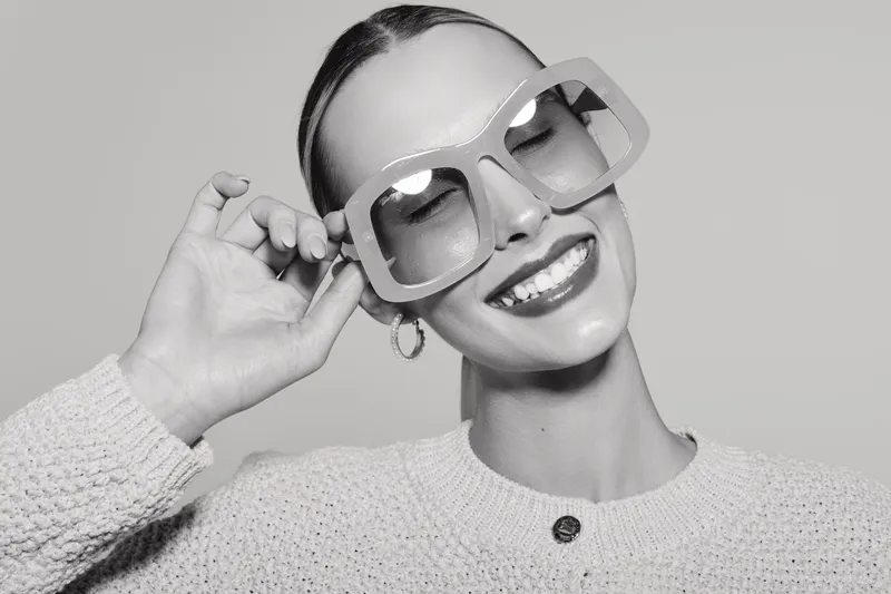 Smiling light skinned woman wearing large sunglasses and knitted sweater, posing with closed eyes and bright smile. Concept of fashion, style, happiness, and accessories.