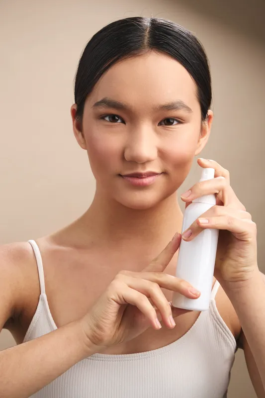 Asian woman with smooth features holds a spray bottle in a bright minimalist bathroom, gazing softly at the camera, conveying freshness, self care, and everyday grooming routine.