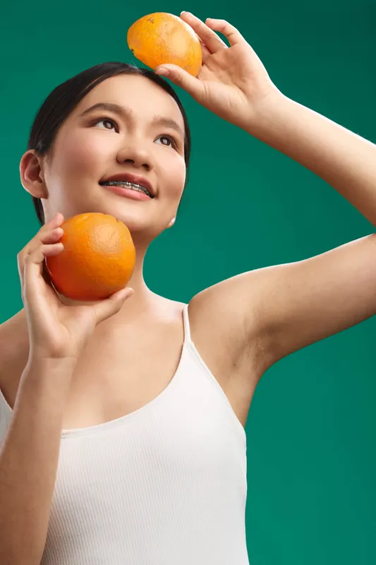 asian woman holding oranges in a bright studio, smiling as she positions fruit against a vivid green backdrop, conveying freshness, vitality, and a playful healthy lifestyle vibe.