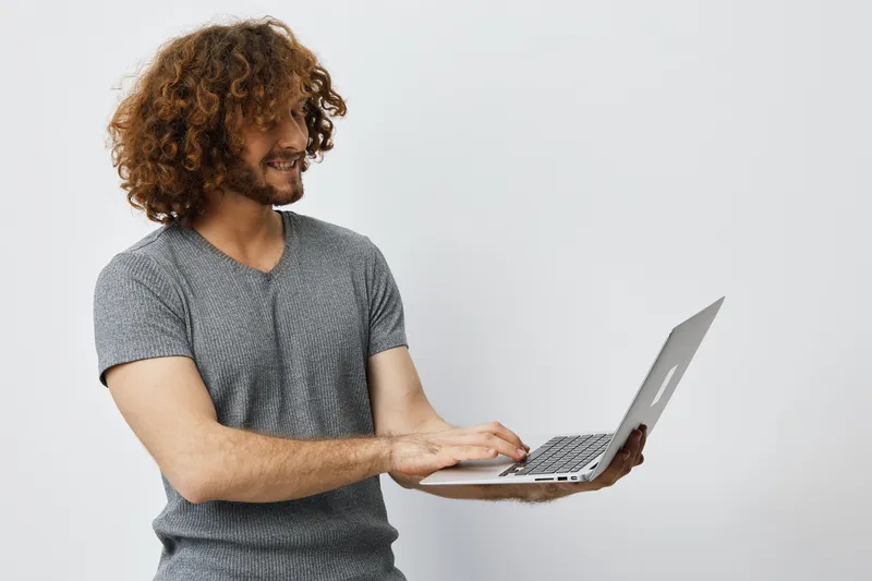 Smiling young man with curly hair using a laptop, wearing a gray shirt, against a light grey background, ideal for concepts of technology and remote work