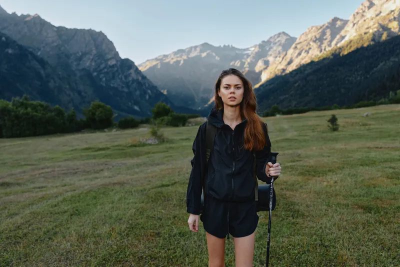 A young woman stands alone in a grassy valley with rugged mountains in the background. She wears a dark outdoor jacket and shorts, holding a trekking pole and looking at the camera with calm