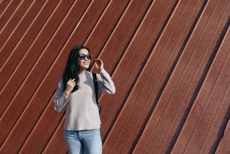 Woman wearing a sweater and jeans with a backpack posing beside a diagonal wooden wall, sunglasses on, casual street style, outdoor fashion and travel vibe for lifestyle and fashion use
