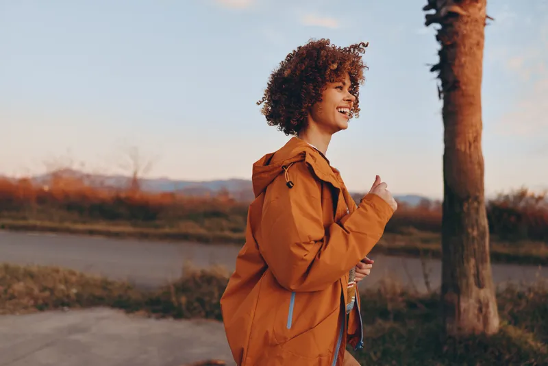 Woman lifestyle outdoors, smiling with curly hair, wearing an orange jacket while walking on a riverside path at golden hour, warm sky, soft breeze, natural colors, relaxed pose, trees