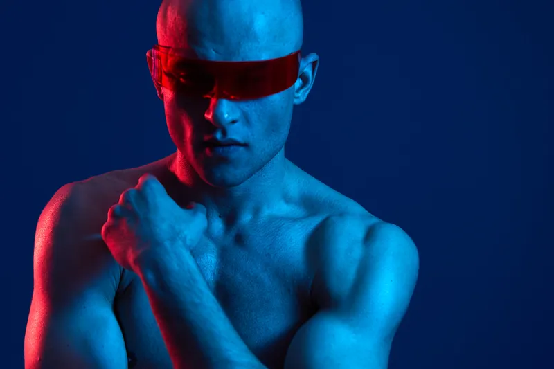 A powerful man with futuristic sunglasses poses dramatically against a vibrant blue background, radiating confidence and strength in a color-light composition.