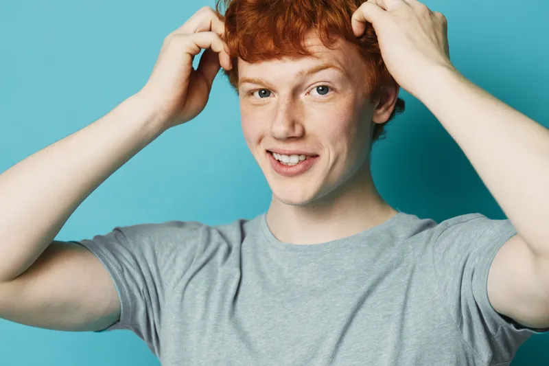Young man with red hair smiling and touching head in casual gray t shirt on blue background. Friendly expression and relaxed pose for portrait or lifestyle concept.