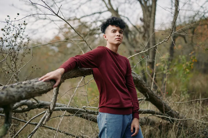 A candid portrait of a young man leaning on a weathered fence in an autumn field, expressing authenticity and credibility through posture, light, and quiet realism.