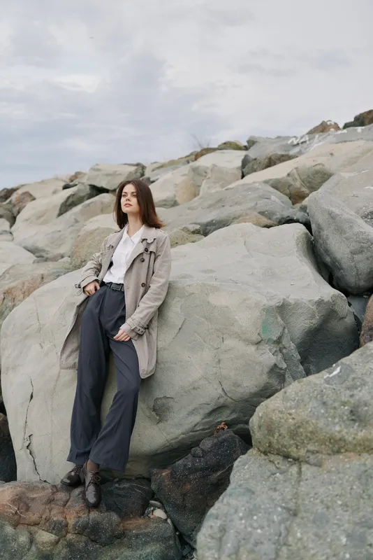 Young woman in stylish outfit stands on rocky shore with cloudy sky background. Fashion concept with natural landscape and casual autumn clothing style outdoors.