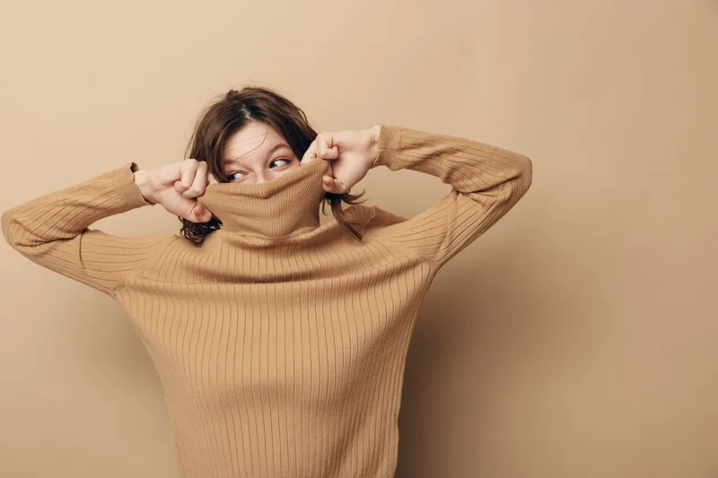 Young woman in beige knitted sweater pulls collar over face, looking sideways with playful expression. Neutral background highlights cozy autumn fashion and casual lifestyle concept.