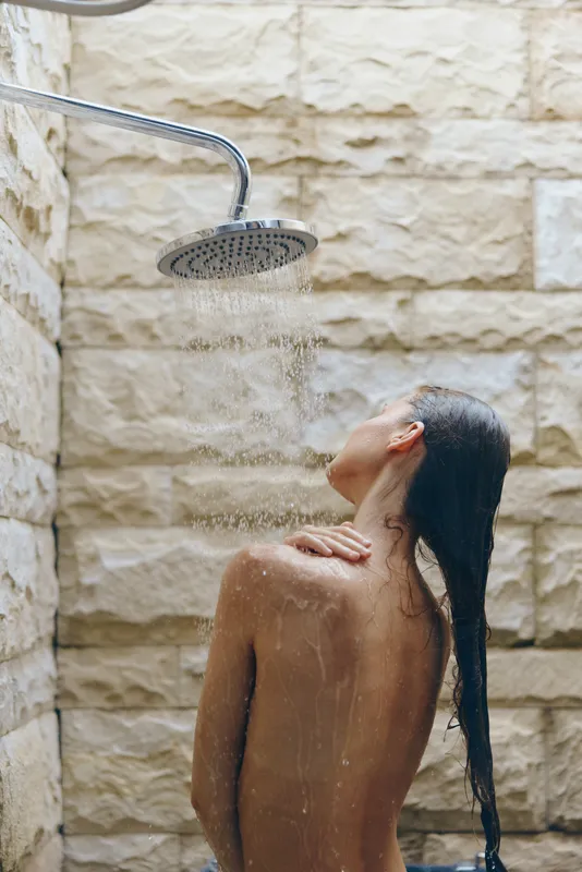 Woman taking outdoor shower with water flowing down back, natural stone background, wet hair, relaxing moment, warm light, summer wellness.