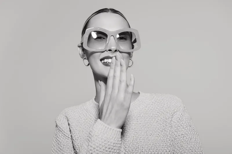 Young white woman with light skin wearing oversized sunglasses giggles holding hand near mouth, isolated on white background.