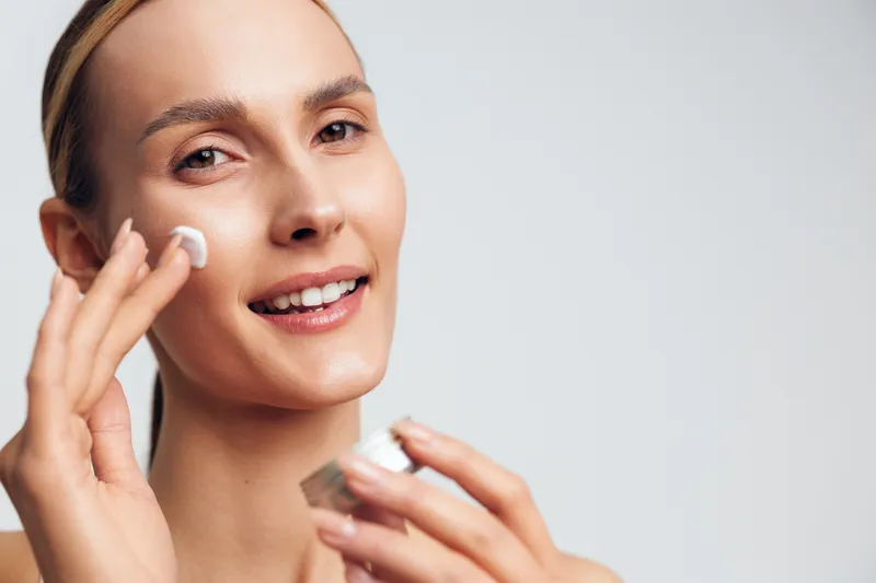 Smiling woman with clear skin applying white face cream on her cheek. Close-up portrait on a light background. Concept of skincare, beauty routine, and self-care with natural light.