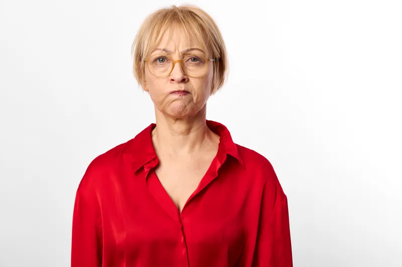 Senior woman with glasses in red blouse, neutral expression, studio portrait on white background, calm mood, close framing, copy space for text, minimalist studio setup