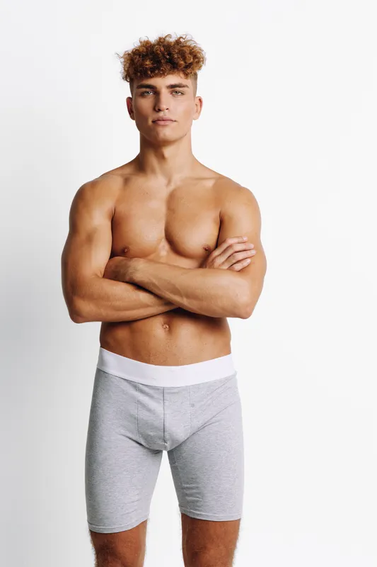 Young muscular man standing with arms crossed wearing gray boxer shorts, looking confidently at camera against white background. Athletic body, curly hair and fitness pose portrait.
