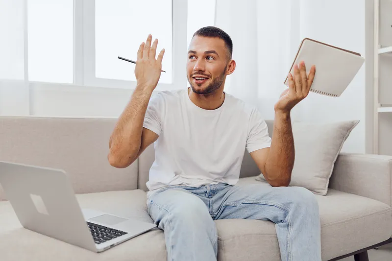 Engaged man explaining ideas while working from home, sitting on a couch with a laptop and notebook, showcasing joy and creativity. Perfect for modern work concepts.