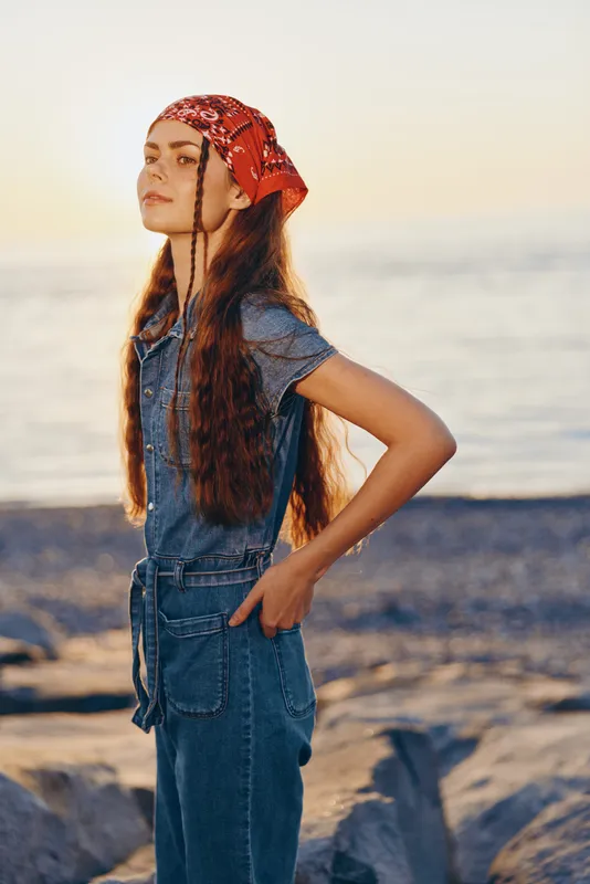 Lifestyle woman wearing modern boho-western denim street style with a red bandana, enjoying golden hour light by the sea in warm film color tones.