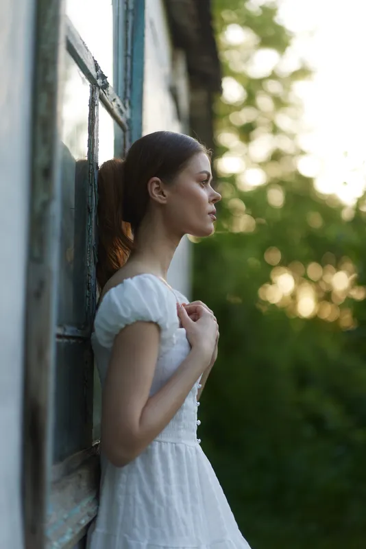Young woman in a white dress, standing outdoors with a peaceful expression, surrounded by green nature at sunset, evoking feelings of tranquility and beauty