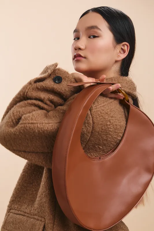 asian woman wearing a soft brown coat holds a curved leather handbag over her shoulder, posing with a confident gaze against a warm backdrop, expressing contemporary fashion and urban chic.
