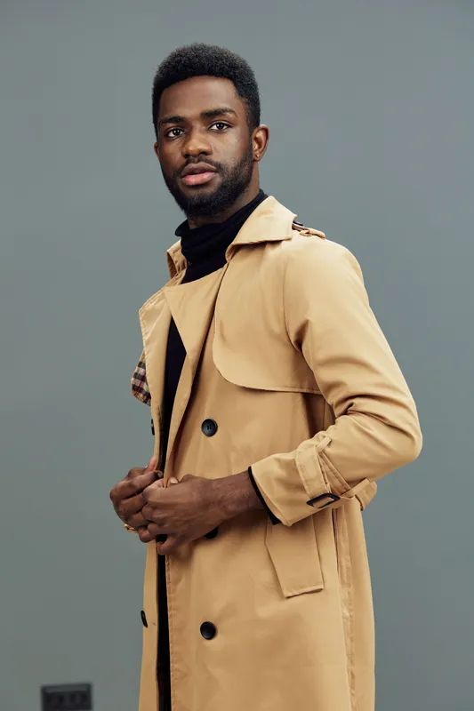 Stylish young Black man wearing a beige trench coat poses confidently against a gray background, showcasing modern fashion and seasonal trends