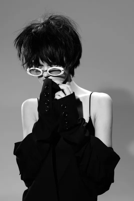 Black and white portrait of a stylish woman wearing white sunglasses and oversized black sleeves expressing shyness and mystery.