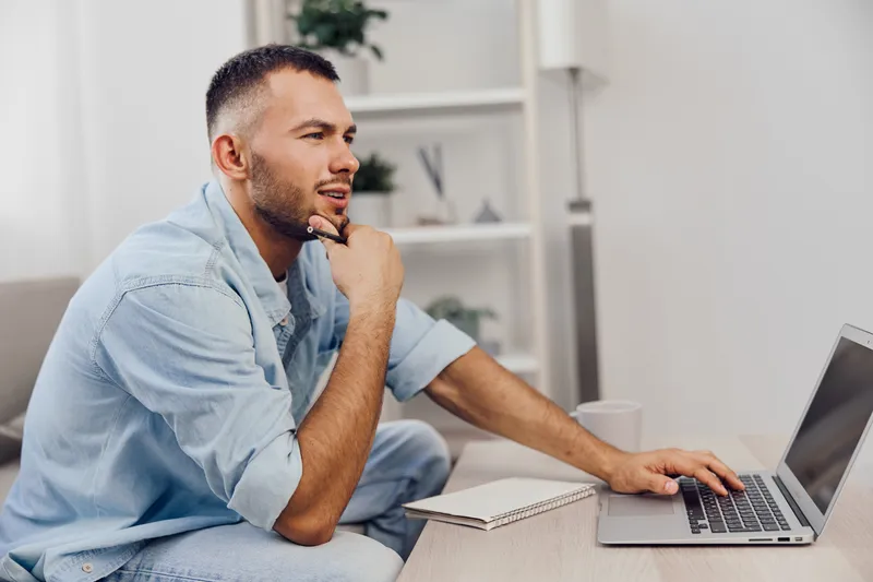 Thoughtful man working on a laptop in a modern home office, focused on his tasks, surrounded by plants and a cozy atmosphere that promotes productivity.