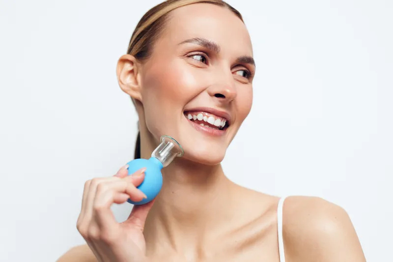 Smiling woman with clear skin holding blue facial massage tool near neck. White background, natural light, concept of skincare, beauty, wellness, and self-care routine for healthy skin.