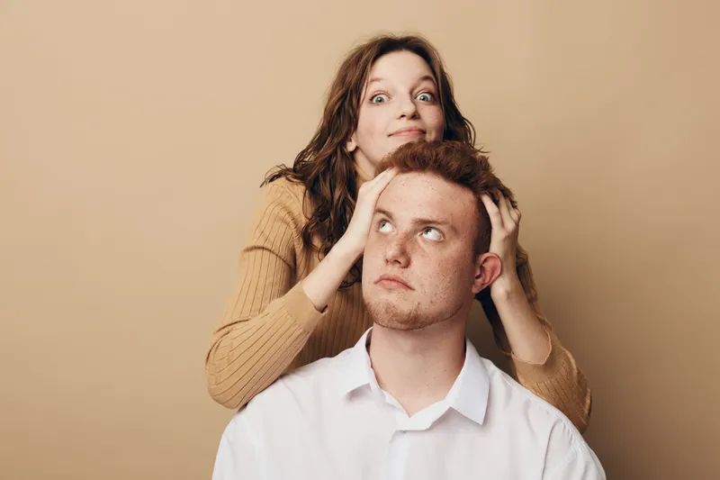 Young woman playfully holds young man’s head from behind on beige background. Casual clothing, funny expression, concept of friendship, humor, and lighthearted moments between friends.