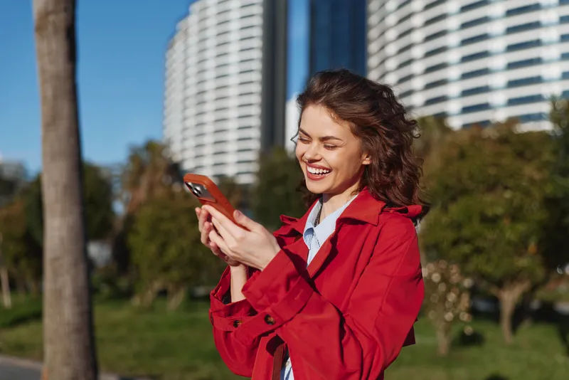 Young woman with curly hair smiles while using a smartphone outdoors on a sunny day with modern buildings and greenery in the background.