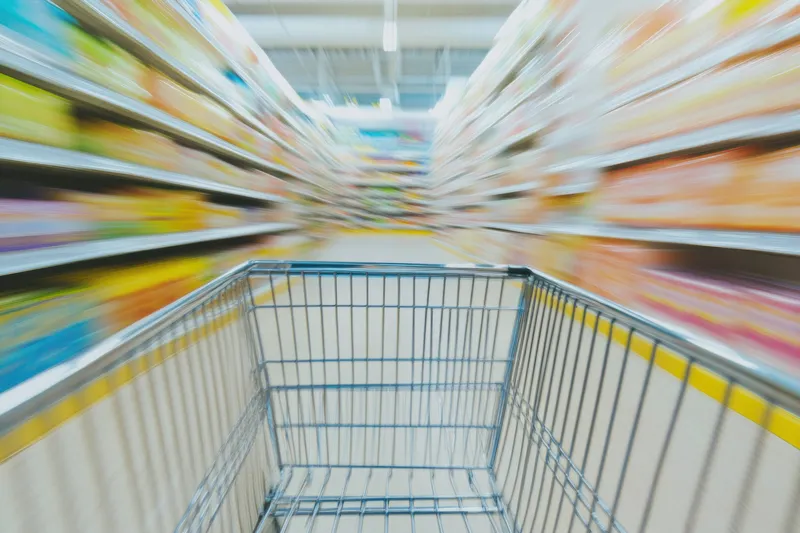Пустая тележка для покупок мчится по яркому проходу продуктового магазина, создавая ощущение движения на фоне размытых полок, заполненных товарами