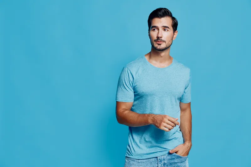 Handsome man in blue t-shirt posing confidently with a thoughtful expression against a solid turquoise background. Stylish and modern lifestyle concept.
