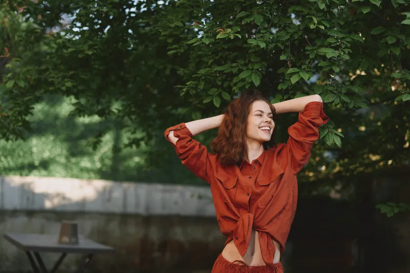 Smiling woman in an orange shirt stands outdoor, arms raised, capturing a portrait of joyful lifestyle amid nature and greenery, with casual fashion and sunny light