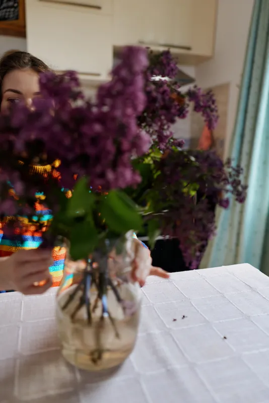A woman sits at a bright kitchen table focused on photographing a vase of purple flowers, the moment conveys quiet warmth and domestic creativity in a home setting.
